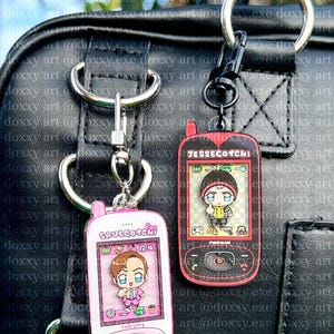 May include: Two acrylic keychains shaped like vintage mobile phones. The left one is pink with the text Saulgotchi and a cartoon character. The right one is black with red accents, the text Jessecotchi, and a cartoon character with a thumbs up.