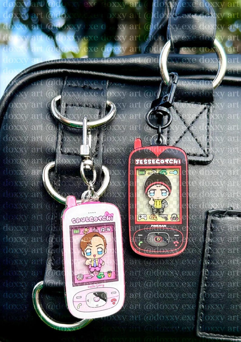 May include: Two acrylic keychains shaped like vintage mobile phones. The left one is pink with the text Saulgotchi and a cartoon character. The right one is black with red accents, the text Jessecotchi, and a cartoon character with a thumbs up.