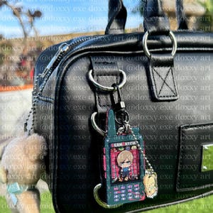 K&ouml;nnte beinhalten: Eine schwarze Handtasche mit einem silbernen Metallring. Ein dunkelgr&uuml;ner, rechteckiger Anh&auml;nger in Form eines alten Mobiltelefons mit der Aufschrift LEON GOTCH ist am Ring befestigt. Ein kleiner, hellbrauner Pl&uuml;schb&auml;r h&auml;ngt daneben.