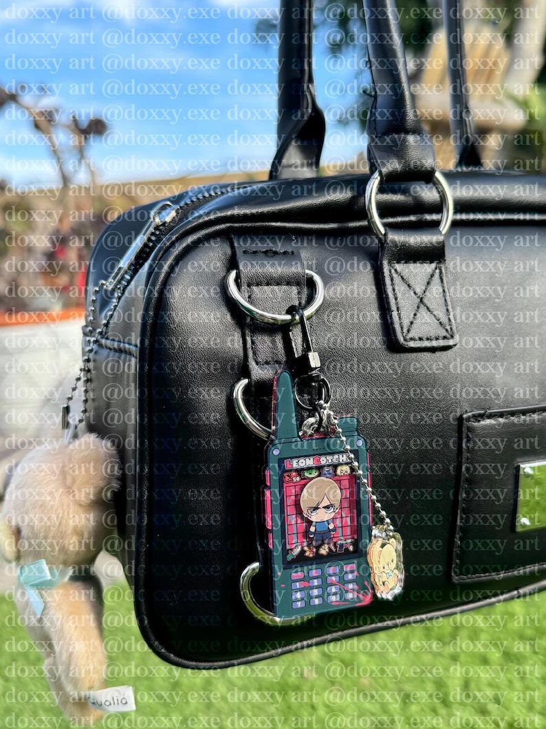 K&ouml;nnte beinhalten: Eine schwarze Handtasche mit einem silbernen Metallring. Ein dunkelgr&uuml;ner, rechteckiger Anh&auml;nger in Form eines alten Mobiltelefons mit der Aufschrift LEON GOTCH ist am Ring befestigt. Ein kleiner, hellbrauner Pl&uuml;schb&auml;r h&auml;ngt daneben.