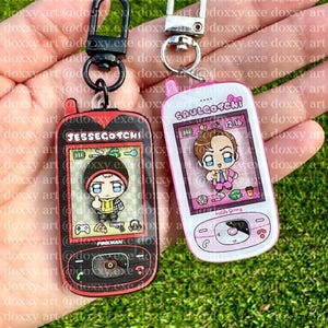 May include: Two acrylic keychains shaped like retro handheld digital pets. The left keychain is black with red accents, labeled JESSEGOTCHI, featuring a cartoon character. The right keychain is white with pink accents, labeled SAULGOTCHI, featuring a cartoon character.