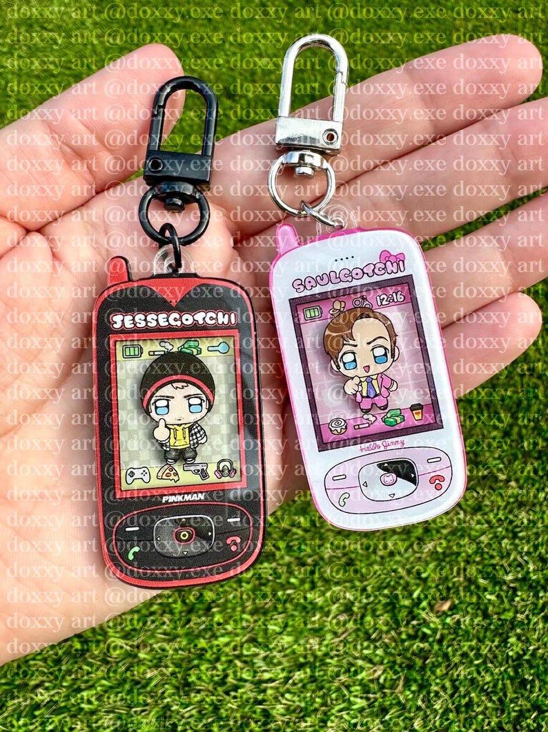 May include: Two acrylic keychains shaped like retro handheld digital pets. The left keychain is black with red accents, labeled JESSEGOTCHI, featuring a cartoon character. The right keychain is white with pink accents, labeled SAULGOTCHI, featuring a cartoon character.