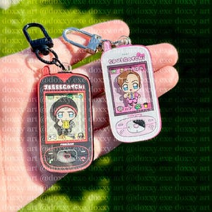 May include: Two acrylic keychains shaped like retro handheld digital pets. The black one features the text JESSEGOTCHI and a cartoon character in a yellow hoodie. The white and pink one features the text SAULGOTCHI and a character in a suit.
