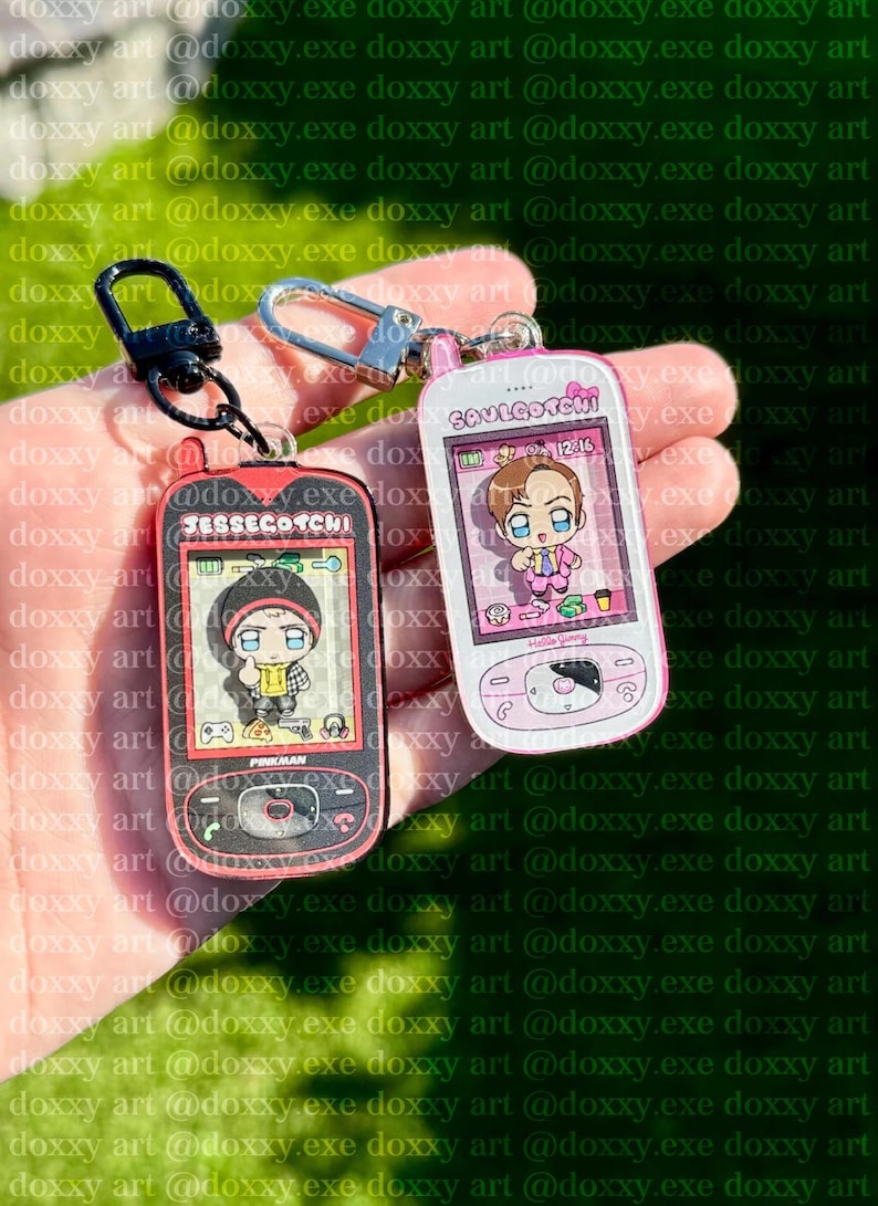 May include: Two acrylic keychains shaped like retro handheld digital pets. The black one features the text JESSEGOTCHI and a cartoon character in a yellow hoodie. The white and pink one features the text SAULGOTCHI and a character in a suit.