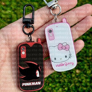 May include: Two rectangular acrylic keychains shaped like vintage mobile phones. One is black with a red Shadow the Hedgehog silhouette and the text PINKMAN. The other is white with a Hello Kitty graphic and the text Hello Jimmy.