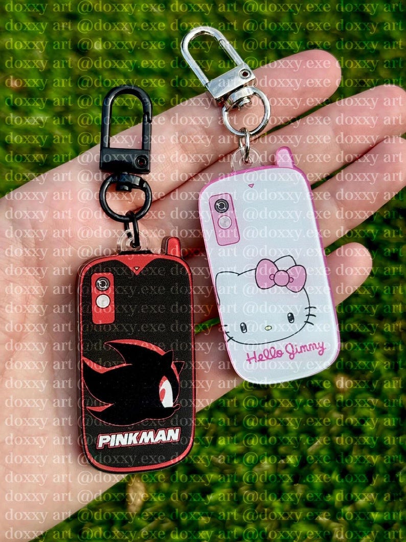 May include: Two rectangular acrylic keychains shaped like vintage mobile phones. One is black with a red Shadow the Hedgehog silhouette and the text PINKMAN. The other is white with a Hello Kitty graphic and the text Hello Jimmy.