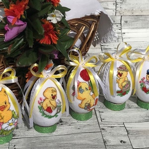 Op de afbeelding: Vijf witte paaseieren versierd met gele strikken en geborduurd met afbeeldingen van kuikens. De eieren worden getoond op een houten oppervlak.