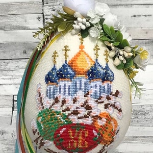 May include: A white Easter egg decorated with a cross-stitch design of a church with blue and gold domes, surrounded by blooming branches and red, green, and orange eggs. The egg is topped with a white flower crown and green leaves.