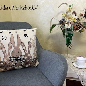 May include: A square cushion with a bunny rabbit design. The cushion features brown bunnies with plaid and polka dot patterns, and a black and white checkered bow. The cushion is on a grey chair.