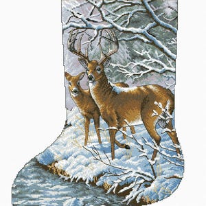 Peut inclure: Un motif de point de croix représentant une scène d'hiver avec deux cerfs debout dans une forêt enneigée au bord d'un ruisseau. Les cerfs sont bruns avec des marques blanches et ont de grands bois. La forêt est recouverte de neige et il y a des branches nues avec de la neige dessus. Le ruisseau est bleu et il y a des rochers dedans.