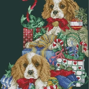 May include: Cross-stitch design showing two Cavalier King Charles Spaniel dogs with brown and white fur, wearing red bows, surrounded by wrapped gifts and festive decorations. The image includes the alphabet in white letters on a dark green background.