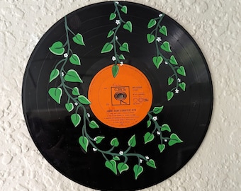 May include: A black vinyl record featuring an orange label with the text "Jimmy Dean's Greatest Hits." The record is decorated with hand-painted green vines, leaves, and small white flowers. The record is mounted on a white wall.