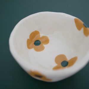 May include: A white ceramic bowl with a floral design. The bowl features three yellow flowers with dark green centres. The bowl is on a dark green background.