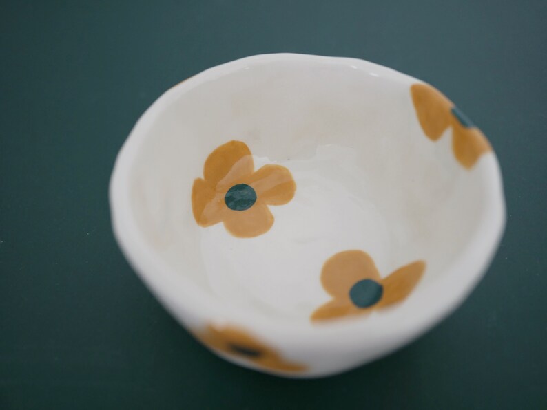 May include: A white ceramic bowl with a floral design. The bowl features three yellow flowers with dark green centres. The bowl is on a dark green background.