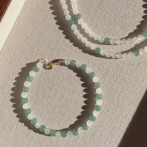 May include: A set of two necklaces made of white and green jade beads. The necklaces are on a light brown fabric surface.