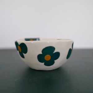 May include: A small white ceramic bowl with a scalloped rim. The bowl is decorated with a repeating pattern of green and yellow flowers.