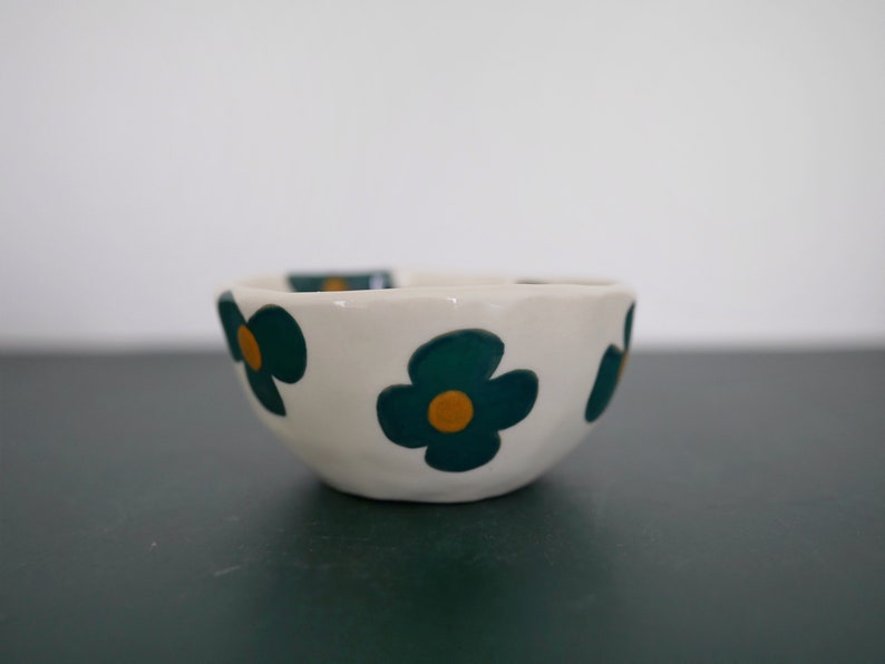 May include: A small white ceramic bowl with a scalloped rim. The bowl is decorated with a repeating pattern of green and yellow flowers.
