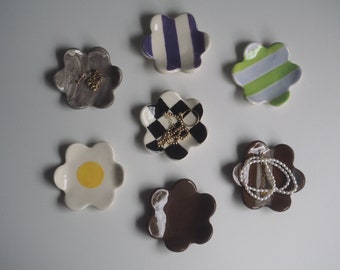 May include: Six ceramic flower-shaped trinket dishes in various colors and patterns. The dishes are arranged on a white surface. Each dish has a different design, including stripes, checkered, and marbled patterns. Some dishes have jewelry on them.