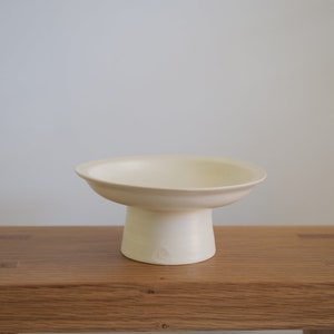 May include: A white ceramic pedestal bowl with a wide, shallow bowl and a round base. The bowl is sitting on a wooden surface.
