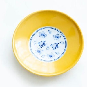 May include: A yellow ceramic saucer with a blue and white floral design featuring two birds. The saucer has a glossy finish.