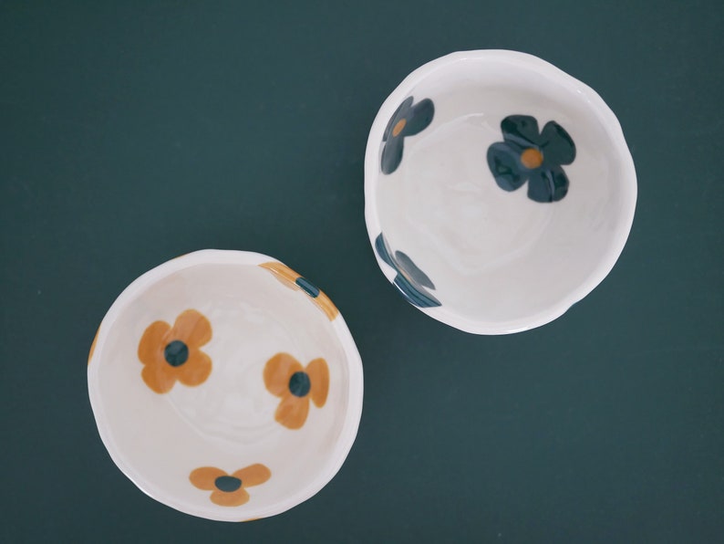 May include: Two white ceramic bowls with floral designs. One bowl has blue and yellow flowers, the other has yellow and black flowers.