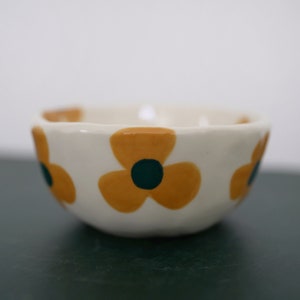 May include: A small white ceramic bowl with a scalloped rim. The bowl is decorated with three yellow flowers with green centres.