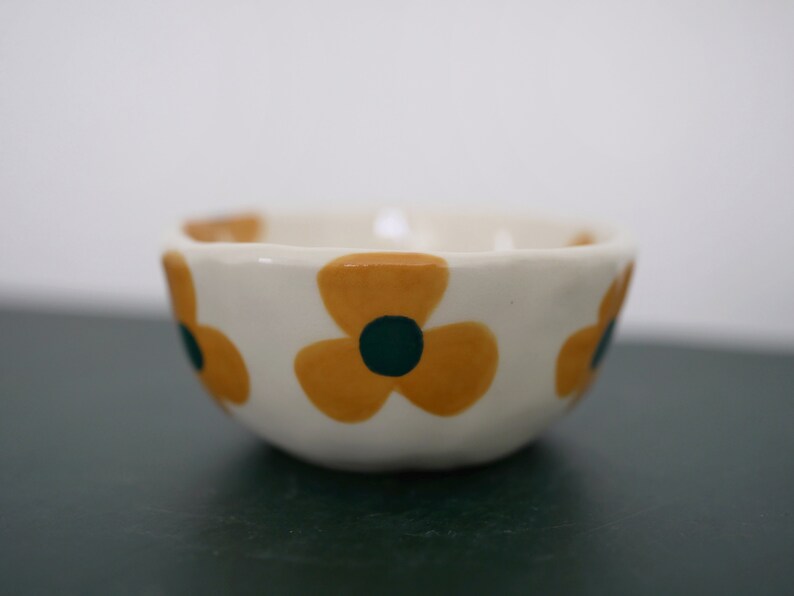 May include: A small white ceramic bowl with a scalloped rim. The bowl is decorated with three yellow flowers with green centres.