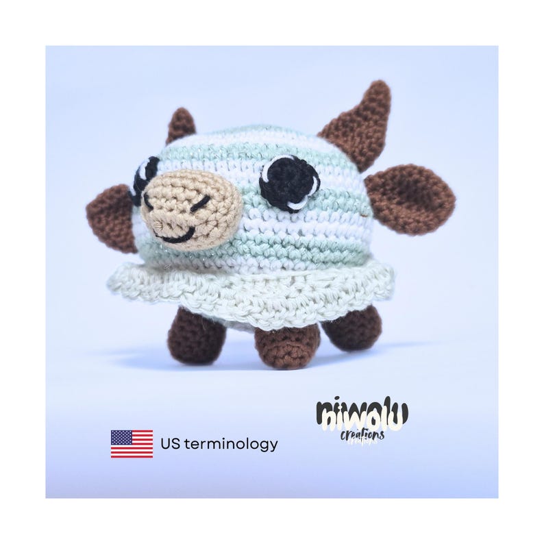 May include: A handmade crochet cow toy with a light blue and white striped head, brown horns and ears, and a beige face with a smiling mouth. The toy has a white frilly skirt and brown legs. The image includes the text "US terminology".