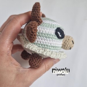 May include: A handmade crochet turtle in shades of green, white, and brown. The turtle has a rounded shell, small legs, and a detailed head with a large black eye. The item is held in a hand.