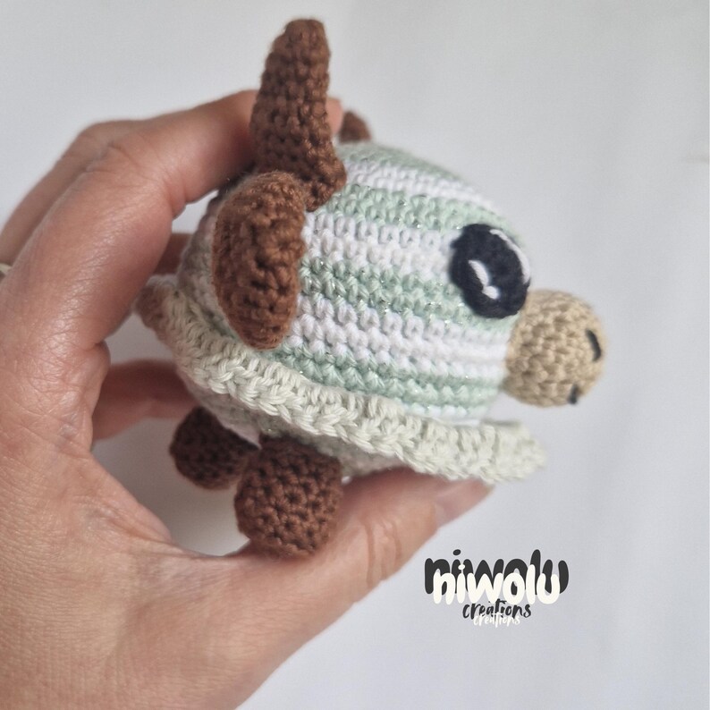 May include: A handmade crochet turtle in shades of green, white, and brown. The turtle has a rounded shell, small legs, and a detailed head with a large black eye. The item is held in a hand.