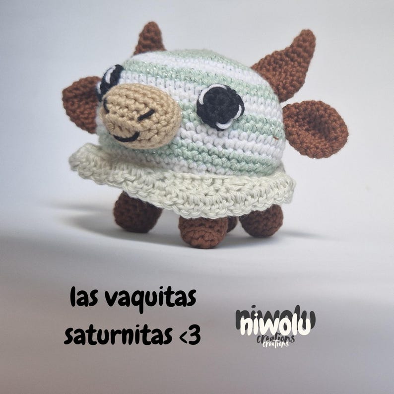May include: A handmade crochet cow figurine with a striped green and white body, brown horns and ears, and a beige face. The cow has black and white eyes and a smiling mouth. The text "las vaquitas saturnitas <3" is below the cow.