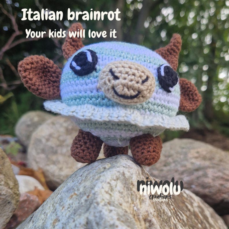 May include: A crocheted creature with a round body, brown horns, ears, and feet, and a smiling face. The body is primarily blue and white with a frilly edge. Text on the image reads "Italian brainrot, Your kids will love it."