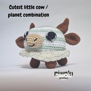 May include: A crocheted cow-planet combination toy. The toy features a light brown face, dark brown ears and legs, and a striped body in white, light green, and silver. The text "Cutest little cow / planet combination" is above the toy.