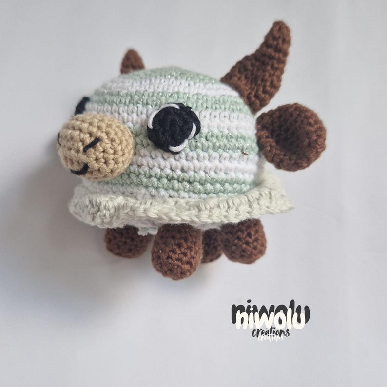 May include: A handmade crochet cow toy with a striped head in white, light green, and silver yarn. The cow has brown horns, ears, and hooves, with a beige muzzle and black and white eyes. The toy is on a white background.