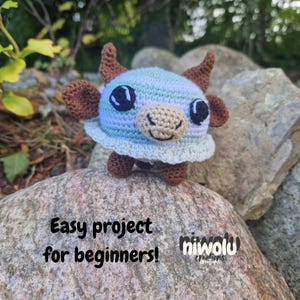 May include: A crocheted blue, brown, and beige cow figurine with large black eyes and small horns. The cow is sitting on a rock. Text on the rock reads "Easy project for beginners!" and the Niwolu Creations logo.