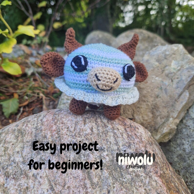 May include: A crocheted blue, brown, and beige cow figurine with large black eyes and small horns. The cow is sitting on a rock. Text on the rock reads "Easy project for beginners!" and the Niwolu Creations logo.