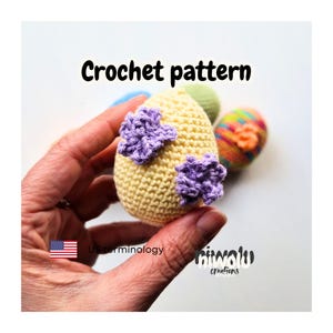 Può includere: Un uovo di Pasqua all'uncinetto giallo chiaro, adornato con due applicazioni floreali viola. L'immagine mostra anche altre uova all'uncinetto colorate. Il testo "Crochet pattern" è visualizzato in alto.