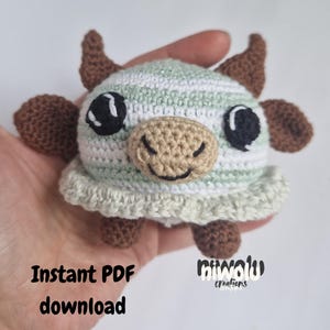 May include: A handmade crochet cow toy with a striped head in white and light green. The cow has brown horns, ears, and feet, with large black eyes and a tan snout. The text "Instant PDF download" is visible.