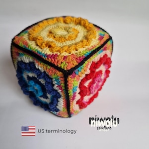 May include: A colorful, crocheted cube with a spiral design on each face. The cube features a variety of colors, including yellow, blue, red, and orange, with black edging. The cube is handmade and has a unique, textured appearance.