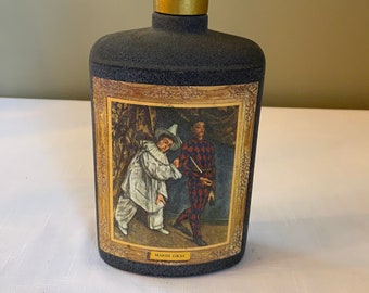 May include: A black ceramic decanter with a gold top and a label featuring a painting of two figures in costumes. The label reads "Mardi Gras".