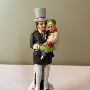 Puede incluir: Una figurita de porcelana de Bob Cratchit y Tiny Tim de Cuento de Navidad. Bob lleva un sombrero de copa y un abrigo marrón, y está sosteniendo a Tiny Tim en sus brazos. Tiny Tim lleva un sombrero verde y un abrigo blanco. La figurita está de pie sobre una base que dice "Bob Cratchit and Tiny Tim".