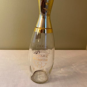 Puede incluir: Una botella de vidrio vintage con forma de pin de bolos con una tapa dorada. La botella tiene una etiqueta que dice "Cream's Pin-Bottle 8 oz."