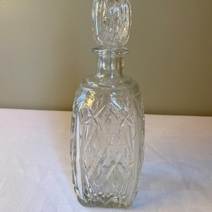 May include: A clear glass decanter with a square body and a diamond-shaped pattern. The decanter has a square stopper with a similar diamond-shaped pattern.
