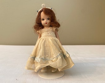May include: A small porcelain doll with brown hair wearing a white dress with blue trim. The doll is standing on a white pedestal.