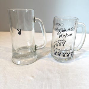 May include: Two clear glass beer mugs with handles. One mug features a black Playboy bunny logo, and the other has the words "Chicago Playboy Club" and additional bunny logos. Both mugs are on a white surface.
