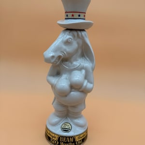 Puede incluir: Botella de cerámica blanca con forma de burro que lleva un sombrero de copa con detalles rojos, blancos y azules. La botella tiene una etiqueta que dice "Beam Bourbon 120 Months Old". Un artículo de colección.