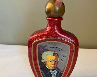 May include: A red glass decanter with a gold stopper and a painted image of Wolfgang Amadeus Mozart. The decanter has a gold trim and the words "W.A. Mozart" are printed on the bottom.