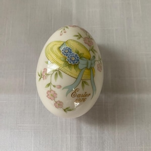 以下が含まれることがあります： 黄色い帽子と青い花が描かれた白い磁器の卵。ピンクの花と緑の葉で飾られています。卵には「Easter 1976」の文字が書かれています。