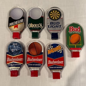 May include: Eight different beer tap handles with various sports themes. The handles are made of clear plastic with a red base. The sports themes include golf, baseball, basketball, football, racing, and darts. The beer brands include Budweiser, Bud Light, Michelob, and O'Doul's.