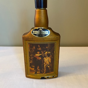 May include: A gold-colored glass bottle with a black plastic stopper. The bottle has a label that says "Beam's Choice 4/5 Quart". The bottle has a picture of a group of people in a painting on the front.